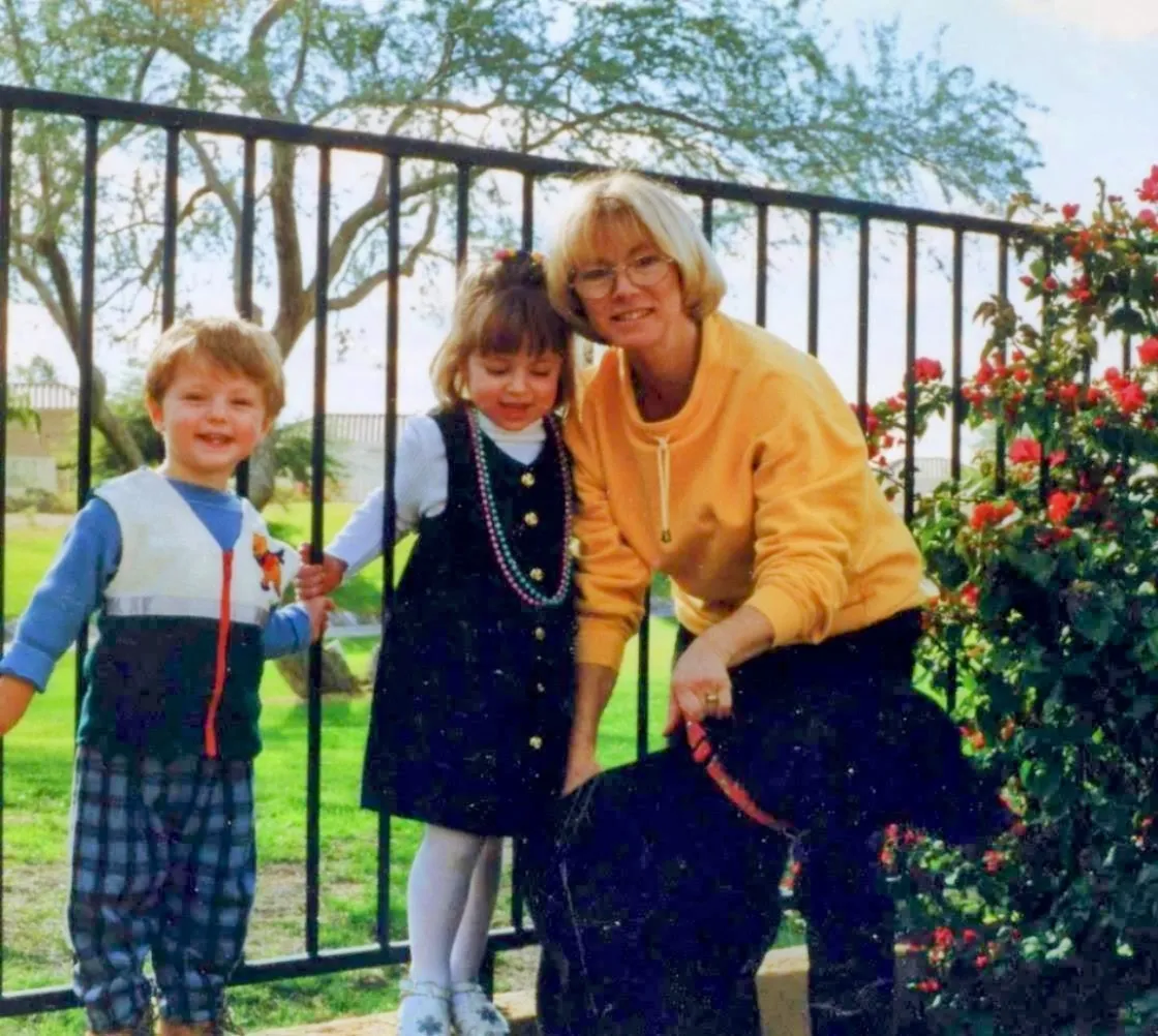 Suzanne with young Zach and Dara and their black dog, beside a fence with roses